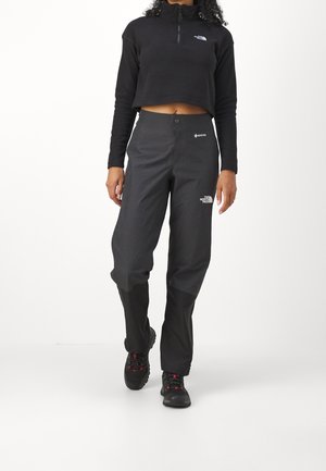 Woman wearing black cropped zip-up fleece and black Gore-Tex hiking pants with rugged black and red boots, walking forward on white background.