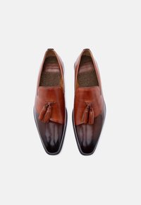 Ζευγάρι λαμπερών καφέ δερμάτινων μοκασίνια με φούντα, σκούρα μύτη και εσωτερικό επενδυμένο με shearling σε λευκό φόντο.