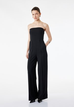 Kvinde iført sort strapless jumpsuit med vide ben og lommer, kombineret med sorte spidse hæle samt sølvfarvede creoler og armbånd.