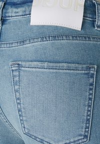 Gros plan sur un jean denim bleu clair, mettant l’accent sur la poche arrière et une étiquette de marque blanche cousue à la ceinture.