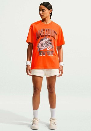Frau trägt ein leuchtend orangefarbenes Nike Court New York T-Shirt, weiße Shorts, Handgelenkbänder und Turnschuhe, steht vor einem einfarbigen Hintergrund.
