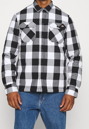 Homme portant une chemise à carreaux noire, blanche et grise avec boutons, deux poches poitrine, et un jean bleu.