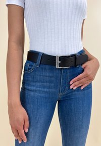 Ceinture en cuir noir avec une boucle rectangulaire en argent, portée sur un jean en denim bleu. Le jean a une coupe classique et une texture subtile.