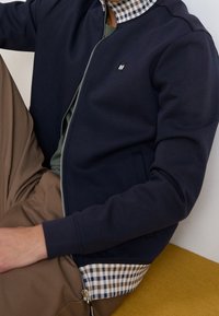 Giacca zip-up blu navy con fodera interna a quadri, caratterizzata da un colletto alto, tasche laterali e una consistenza liscia. Indossata sopra un top verde.