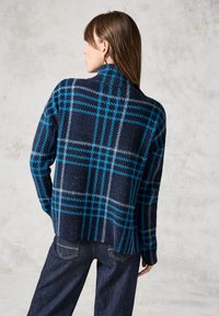 Merino-punottu villapaita tummansinisellä ruudullisella kuviolla, jossa on korkea kaulus ja rento malli, yhdistettynä tummiin farkkuihin.