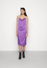 Vestido slip de cetim roxo com decote em cowl e alças finas, apresentando fendas laterais, combinado com botins pretos de bico fino.
