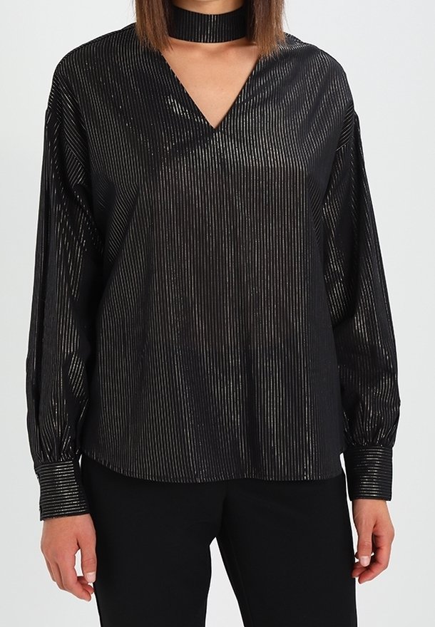 Femme portant un chemisier noir brillant rayé à manches longues avec un col en V et un collier assorti, associé à un pantalon noir.