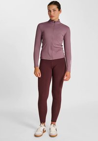 Top met lange mouwen en ritssluiting in lichtpaars, gecombineerd met bordeauxrode leggings. Model draagt witte en bruine sneakers. Soepele, aansluitende stof.