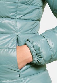 Hand insatt i den dragkedjade fickan på en ljusblå vadderad jacka med elastisk mudd på ärmen.