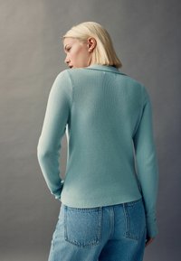 Haut à manches longues bleu clair à côtes avec un col, associé à un jean en denim à taille haute de lavage moyen. Texture lisse, lignes épurées.