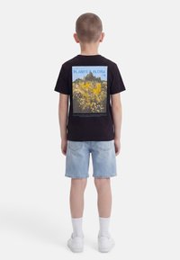 Αγόρι που φοράει μαύρο T-shirt με μεγάλο έγχρωμο σχέδιο βουνού και λουλουδιών στην πλάτη, τζιν σορτς, λευκές κάλτσες και λευκά αθλητικά παπούτσια, όρθιο.