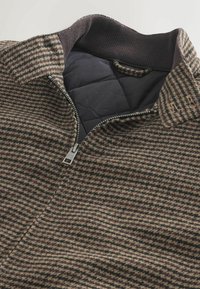 Rudai ir tan houndstooth rašto striukė su užtrauktuku, briaunotu apykakle ir paminkštinta juoda vidine dalimi. Struktūrizuota vilnos mišinio medžiaga.