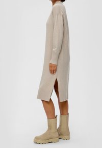 Abito lavorato a maglia beige con maniche lunghe, spacco laterale e orlo a coste. Abbinato a stivaletti beige alla caviglia con suola spessa e chiusura con zip.