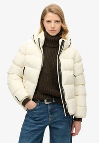 Giacca imbottita bianca con cappuccio, dotata di una cerniera nera e dettagli in nero. Indossata sopra un maglione di lana marrone e jeans blu.