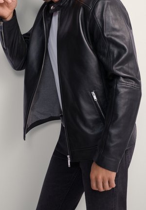 Mężczyzna ubrany w czarną skórzaną kurtkę na białej koszuli z czarnymi spodniami, kurtka częściowo rozpięta, ręka swobodnie luźno u boku.