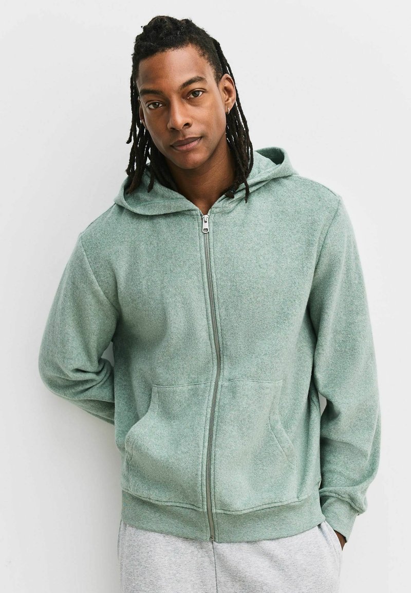 Bărbat cu dreadlocks, purtând un hanorac verde deschis cu fermoar și pantaloni gri deschis, stând cu o mână la spate.