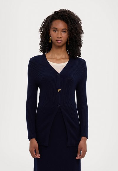 Cardigan a coste di colore blu navy con maniche lunghe, scollo a v e chiusura con un singolo bottone. Presenta un distintivo accento triangolare dorato.