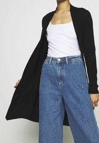 Cardigan negro de punto con frente abierto, mangas largas y textura ligera. Combinado con jeans de mezclilla azul de cintura alta que cuentan con cierre de botón y cremallera.