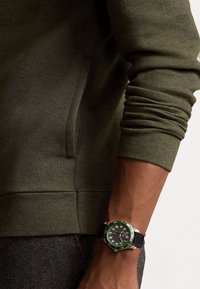 Grüner Pullover aus strukturiertem Stoff und eine Uhr mit grünem bezel, schwarzem Armband und auffälligen weißen Ziffern auf dem Zifferblatt.