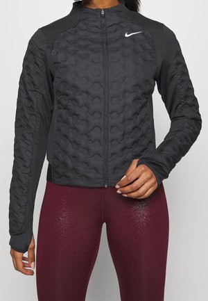 Vrouw draagt een zwarte doorgestikte jas met ritssluiting en Nike-logo en glanzende kastanjebruine leggings, hand past de zoom van de jas aan.