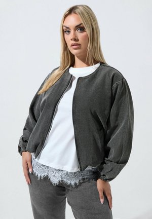 Femme aux longs cheveux blonds porte une veste grise zippée par-dessus une chemise blanche avec bordure en dentelle, debout devant un fond clair uni.