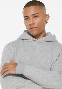 Gray hooded sweatshirt made of soft fabric. Features a kangaroo pocket, ribbed cuffs, and a relaxed fit. Model posing with arms crossed.