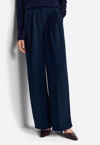 Pantaloni a gamba larga blu navy con pieghe frontali e una texture liscia. Abbinati a scarpe con punta affusolata in una tonalità scura, che mettono in risalto un look elegante.