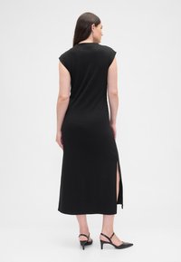 Czarna, długa do ziemi sukienka z krótkimi rękawkami typu cap sleeve, z bocznym rozcięciem i gładką, elastyczną fakturą. Do kompletu czarne sandały na obcasie.