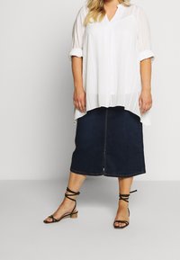 Blusa bianca a righe con scollo a V e maniche arrotolate abbinata a una gonna midi in denim scuro. Indossata con sandali neri con cinturino.