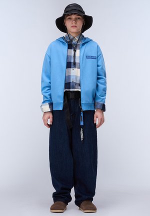 Ung person iført en sort bucket hat, blå hættejakke med lynlås, ternet skjorte, brede mørke jeans og brune sko, stående mod en ensfarvet baggrund.