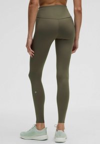 Spodnie legginsy z wysokim stanem w kolorze oliwkowym, wykonane z elastycznego materiału, z gładką teksturą i małym logo w pobliżu kostki. Połączone z jasnooliwkowymi butami.