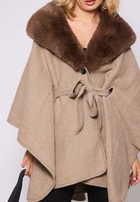 Femme portant une cape en laine beige avec un large col en fourrure brune et une ceinture nouée, tenant un sac à main noir, se tenant devant un fond uni.