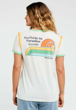 Frau mit welligem Haar, die ein weißes T-Shirt mit grünem Besatz und Retro-Aufdruck „Surfside in Paradise“, Regenbogenstreifen und orangefarbener Sonnen-Grafik trägt.