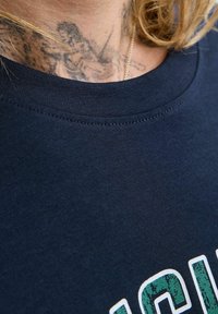 Nakketatovering af en figur, der holder en bue og pil, delvist dækket afblond hår og en marineblå skjorte med grønne og hvide bogstaver.