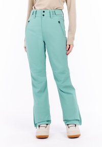 Vrouw in turquoise waterdichte sneeuwbroek met ritszakken, beige lange mouwtop en witte winterlaarzen voor een witte achtergrond.