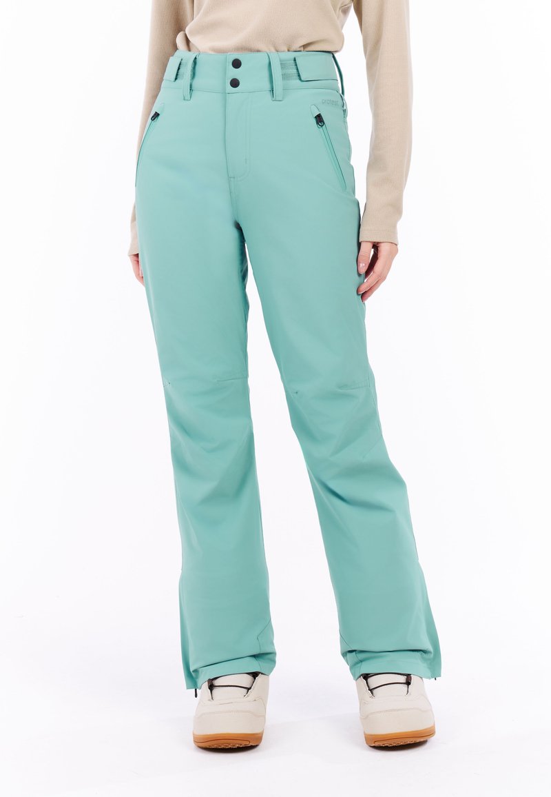 Vrouw in turquoise waterdichte sneeuwbroek met ritszakken, beige lange mouwtop en witte winterlaarzen voor een witte achtergrond.