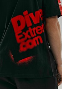 Czarny bawełniany T-shirt z wyrazistym, czerwonym tekstem „DiveExtreme.com” nas sprayem, z efektem postarzania. Krótkie rękawy i luźny krój.
