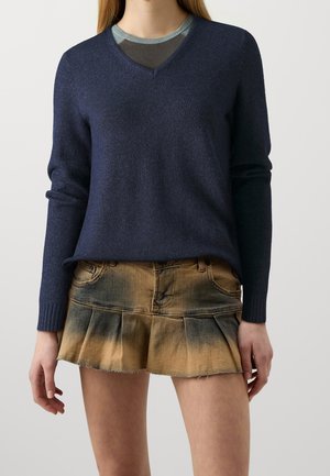 Kobieta ubrana w granatowy sweter z dekoltem w serek oraz krótką, plisowaną spódnicę dżinsową z efektem prania w kolorach brązowym i czarnym.