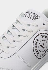 Λευκό αθλητικό παπούτσι με λευκά κορδόνια, κείμενο "Versace Jeans Couture" στη γλώσσα και κυκλικό λογότυπο στο πλάι.