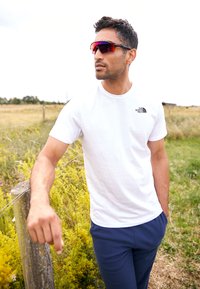 Maglietta bianca a maniche corte con logo nero, pantaloni blu navy e occhiali da sole con tonalità rossa. Ambientazione all'aperto con erba verde e fiori gialli.