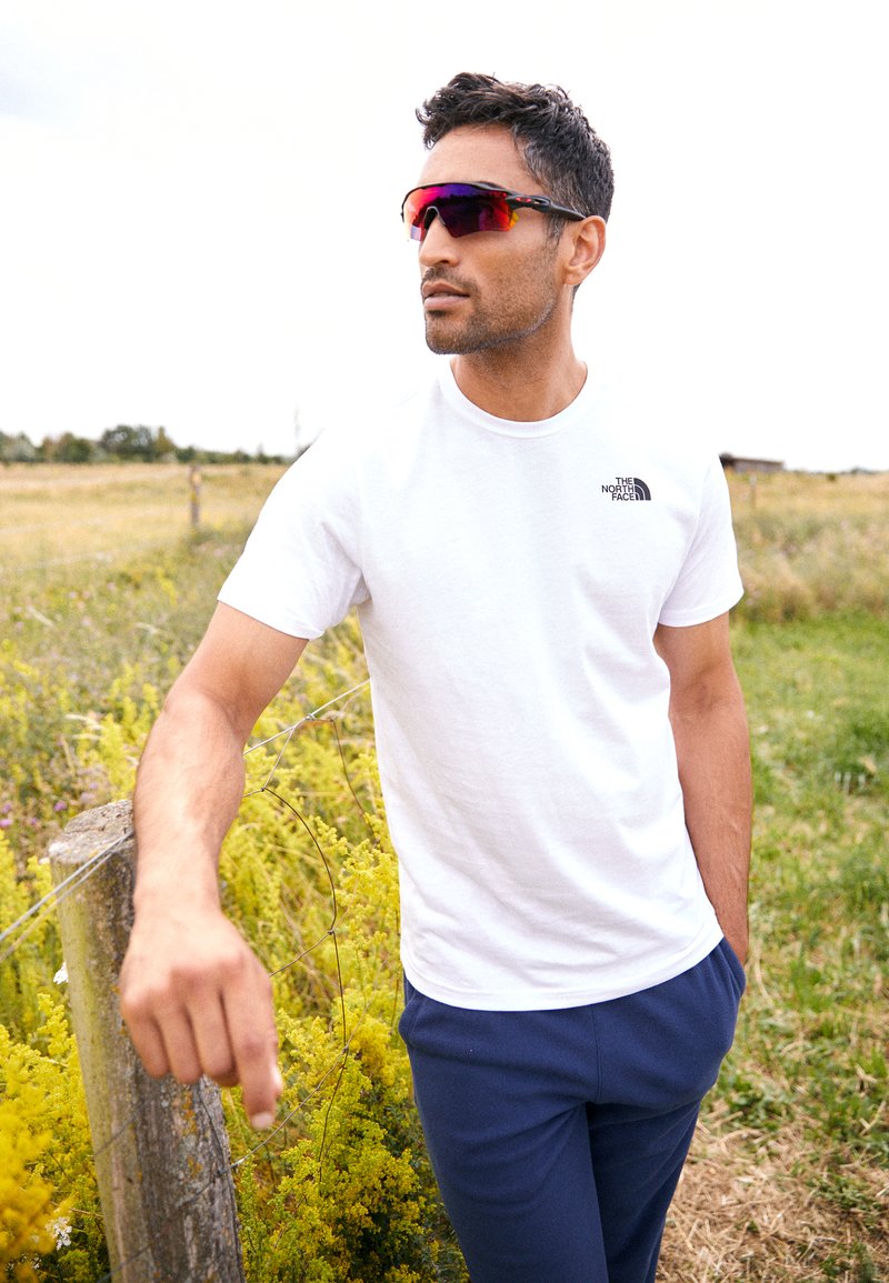 Maglietta bianca a maniche corte con logo nero, pantaloni blu navy e occhiali da sole con tonalità rossa. Ambientazione all'aperto con erba verde e fiori gialli.