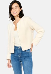 Crème-kleurige, gestructureerde cropped cardigan met ronde hals en twee knopen aan de voorkant, gedragen over een witte top en denim jeans.