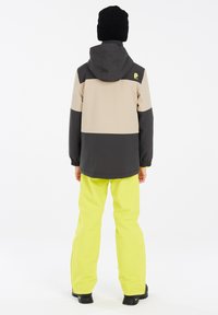 Skijacke mit einem Farbblockdesign in Schwarz und Beige, ausgestattet mit einer Kapuze und elastischen Bündchen, kombiniert mit leuchtend gelben Schneehosen und schwarzen Stiefeln.
