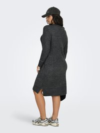 Kvinna klädd i en mörkgrå lång tröjklänning med sidospalt, svart keps och vita sneakers, stående mot en enkel ljus bakgrund.