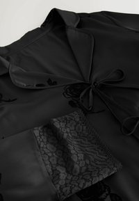 Schwarze Bluse mit Satin-Finish, floraler Struktur und Spitzendetails an den Ärmeln. Verfügt über einen Kragen und eine Bindung zum Verschließen.