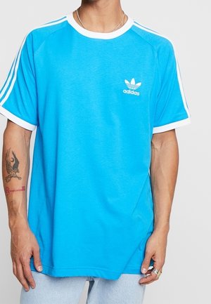 Man met felblauw Adidas-T-shirt met witte bies en strepen op de schouders, lichtblauwe spijkerbroek en een tatoeage op de linker onderarm.