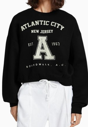 Black cropped sweatshirt with loose sleeves, featuring "ATLANTIC CITY NEW JERSEY" and a textured "A" logo in beige. White drawstring at the hem.