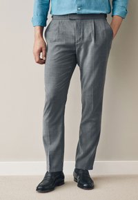 Pantalon habillé gris plissé avec une coupe ajustée, fabriqué dans un tissu texturé. Porté avec des chaussures en cuir noir, accompagné d'une chemise bleue.
