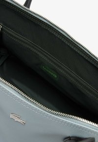Interno di borsa azzurro chiaro con tasca con cerniera, rivestimento nero e piccolo logo Lacoste visibile di lato.