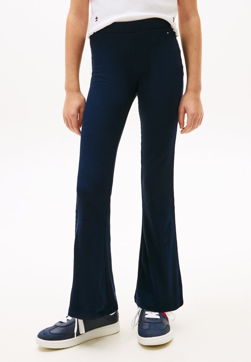 Άτομο που φοράει μπλε σκούρο παντελόνι με ριγέ υφή και φαρδιά παντελόνια και μπλε αθλητικά παπούτσια με άσπρες σόλες και κορδόνια, όρθιο σε λευκό φόντο.